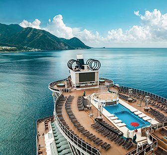 A cruise ship deck featuring a pool and sun loungers with mountains in the background | MSC Cruises