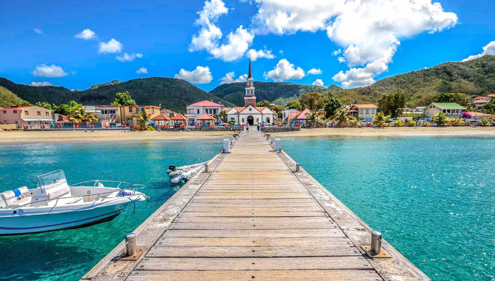 A wooden pier leading to a scenic coastal village with a colorful church and clear turquoise waters | MSC Cruises A wooden pier leading to a scenic coastal village with a colorful church and clear turquoise waters | MSC Cruises